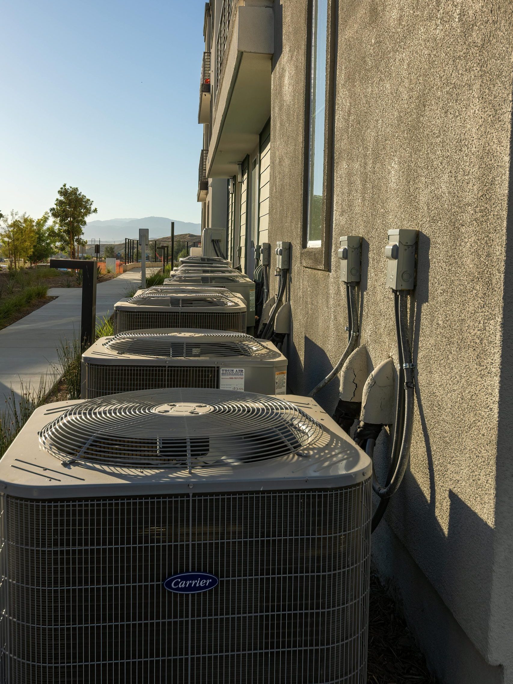 Line of air conditioning units on a city apartment exterior with a sidewalk view.